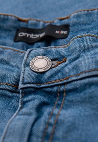Jean en denim bleu avec coutures visibles, featuring a metal button with the brand name "Ombre" and a black tag at the waistband.