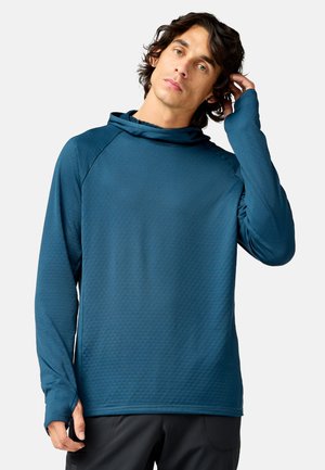 Junger Mann mit dunklem lockigem Haar, der ein blaues langärmeliges Kapuzenshirt mit Daumenlöchern trägt und vor einem schlichten hellen Hintergrund steht.