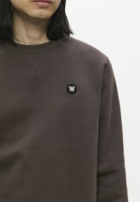 Bruine sweatshirt met een ronde zwarte patch met een witte "W" logo, gladde textuur, ronde hals en zichtbare stikdetails.