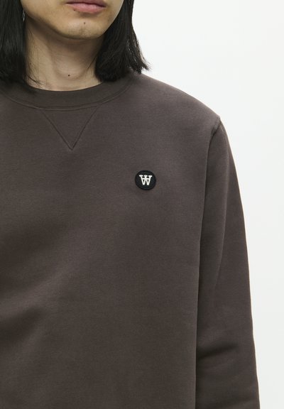 Sweat-shirt marron avec un patch noir rond arborant un logo "W" blanc, texture lisse, col rond et détails de couture visibles.