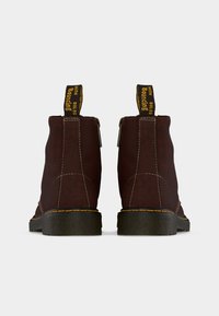 Ζευγάρι σκούρα καφέ δερμάτινων μπότες με μαύρες σόλες και κίτρινα ράμματα, εμφανιζόμενες από πίσω με ταινίες έλξης που γράφουν «WITH BOUNCING SOLES».