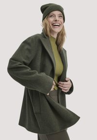 Grüner Mantel aus Wollmischung mit maßgeschneidertem Design, weiten Ärmeln und Seitentaschen, kombiniert mit einem gerippten grünen Oberteil und einer olivfarbenen Mütze.