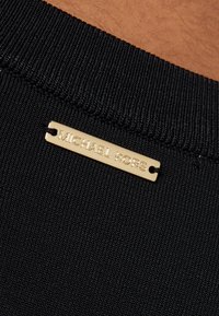 Zwart geribd stof met een gouden metalen label gegraveerd met "MICHAEL KORS," bevestigd aan de halslijn, met een strak, minimalistisch ontwerp.