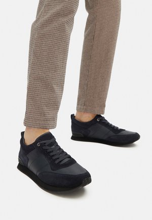 Mornarsko modri športni čevlji z mešanico usnja in gladkega tekstila, s katerimi so zavezovanje na vezalke in gumijastim podplatom, kombinirani s karo hlačami.