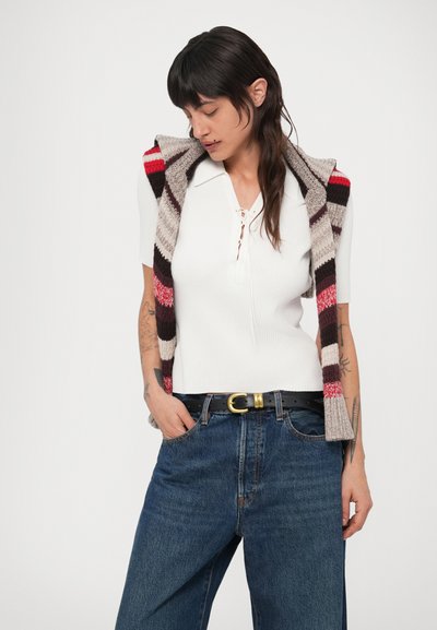 Hosszú sötét hajú nő fehér rövid ujjú kötött felsőben, farmert fekete övvel, és vállára terített csíkos kötött pulóverrel.