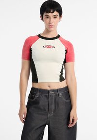 Short-sleeve cropped shirt in off-white, olive green, and pink with ribbed neckline. Features "GUESS" logo on chest. Paired with dark denim jeans.