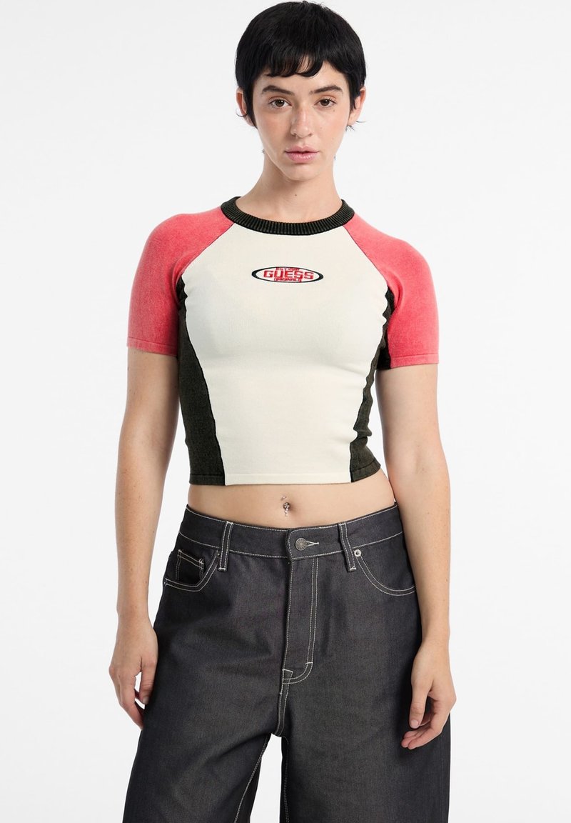 Short-sleeve cropped shirt in off-white, olive green, and pink with ribbed neckline. Features "GUESS" logo on chest. Paired with dark denim jeans.