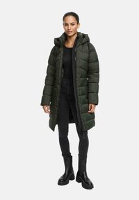 Vrouw gekleed in een lange donkergroene puffercoat met capuchon, zwarte top, zwarte skinny jeans en zwarte chunky boots, staand voor een witte achtergrond.