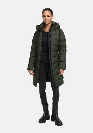 Vrouw gekleed in een lange donkergroene puffercoat met capuchon, zwarte top, zwarte skinny jeans en zwarte chunky boots, staand voor een witte achtergrond.
