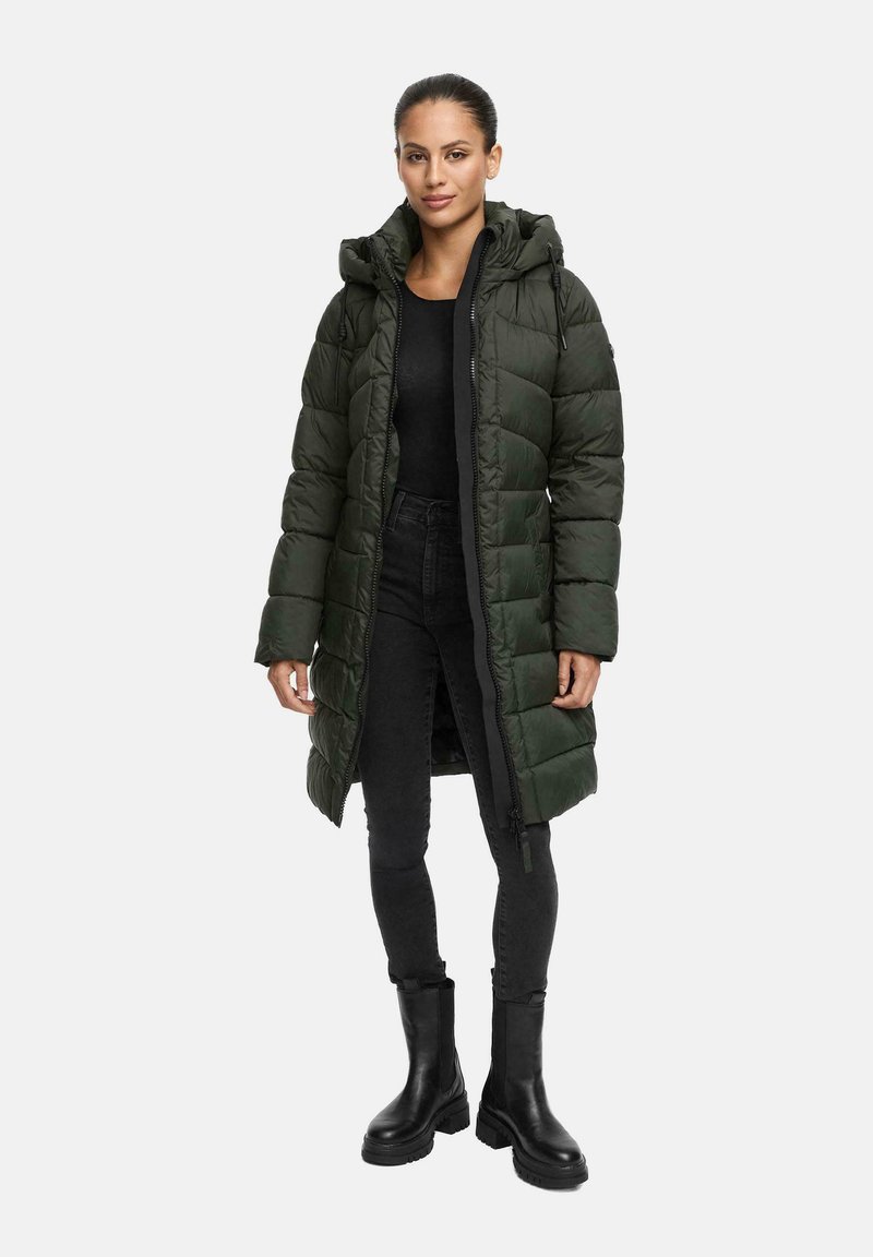 Vrouw gekleed in een lange donkergroene puffercoat met capuchon, zwarte top, zwarte skinny jeans en zwarte chunky boots, staand voor een witte achtergrond.