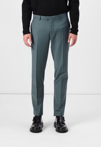 Pantalones en un color teal apagado, hechos de una tela suave, con pliegues frontales y un corte ajustado, combinados con zapatos negros.
