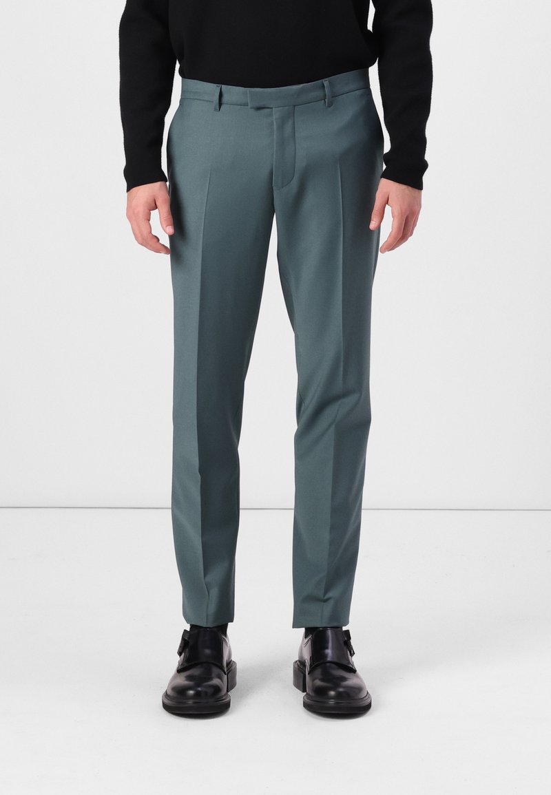 Pantalones en un color teal apagado, hechos de una tela suave, con pliegues frontales y un corte ajustado, combinados con zapatos negros.