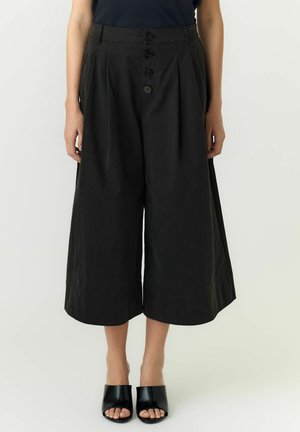 Zwarte wijde cropped broek met knopen aan de voorkant, gedragen met zwarte open-toe mulehakken op een lichte achtergrond.