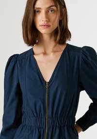 Ολόσωμη φόρμα σε ναυτικό μπλε χρώμα με βαθύ ντεκολτέ V, λεπτομέρεια με χρυστό φερμουάρ, ελαστική μέση και φουσκωτά μανίκια, κατασκευασμένη από λείο ύφασμα.