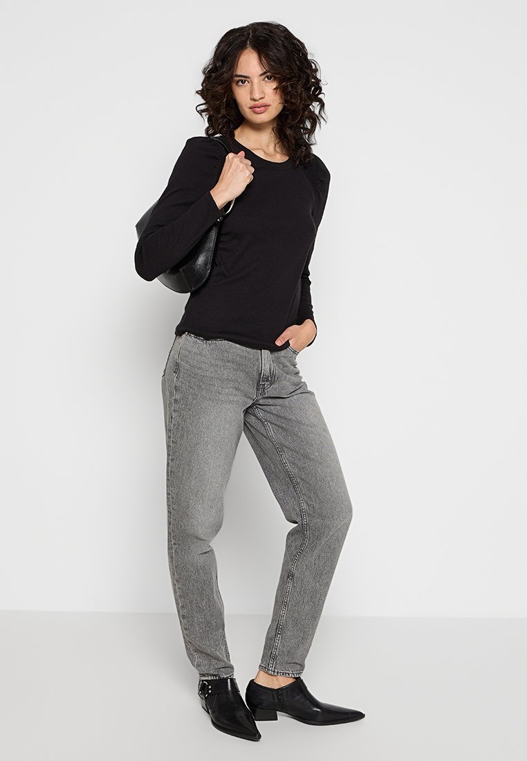 Czarna bluzka z długim rękawem, szare lekko luźne dżinsy z wyblakłym wykończeniem oraz czarne botki za kostkę. Modelka trzyma czarną torbę na ramię na jednolitym tle.