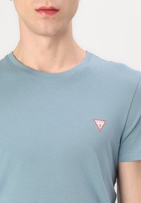 Világoskék, rövidujjú pamut póló, kerek nyakkivágással. Bal mellrészén található egy kis piros háromszög logó, amelyen a "GUESS?" felirat szerepel.
