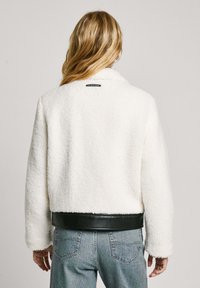 White fluffy jacket made of textured material with a black leather waistband. Features a collar and small brand label at the back.