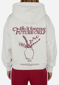 Biely kapucňový sveter s marhuľovým textom "Rob to pre svoj BUDÚCI JA" nad líniovou kresbou vázy s rastlinami.