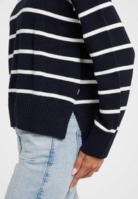 Pulover bleumarin cu dungi albe orizontale, cu tiv și manșete ribbate, purtat cu blugi denim de culoare albastru deschis, cu detalii laterale vizibile.
