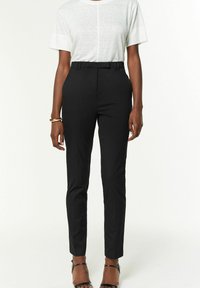 Černé kalhoty na míru se slim fit střihem, vysokým pasem a poutky na opasek, v kombinaci se světle šedým tričkem s krátkým rukávem. Hladká textura látky.