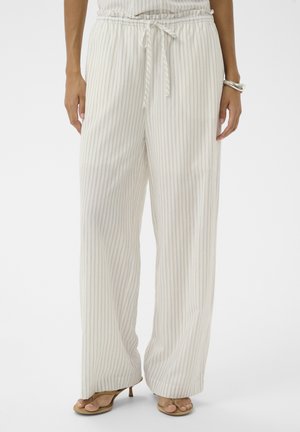Femme portant un pantalon crème taille haute à jambes larges avec de fines rayures verticales et une ceinture nouée, assorti à des talons ouverts beige et un bracelet argenté.