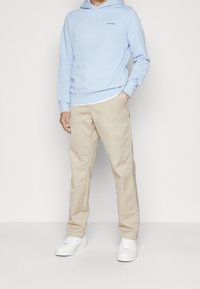 Felpa con cappuccio di colore azzurro chiaro con un piccolo logo, abbinata a pantaloni beige e sneaker bianche su uno sfondo neutro. Texture del tessuto liscia.