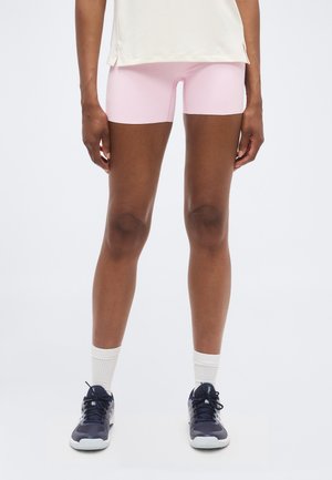 Person iført lyserøde træningsshorts, hvide sokker og sorte sneakers, der står mod en hvid baggrund.