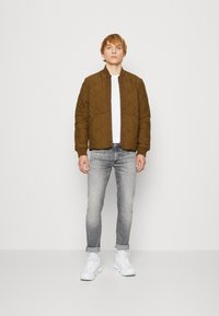 Chaqueta bomber acolchada de color verde oliva con patrón de diamante, puños acanalados, sobre una camiseta blanca y vaqueros gris claro, combinada con zapatillas blancas.