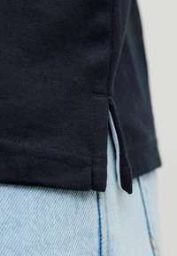 Zbliżenie na rąbek czarnej koszuli z rozcięciem po bokach, noszonej na jasnoniebieskich dżinsach.