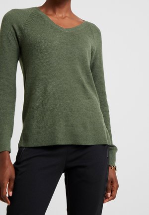Woman wearing a textured olive green V-neck sweater and black pants, with a gold bracelet on her left wrist against a plain background.
