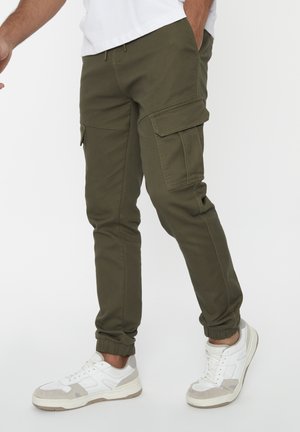 Groene cargobroek met elastische boorden, voorzien van zijzakken en een trekkoord in de taille. Gecombineerd met witte sneakers en een effen shirt.