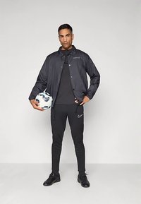 Hombre con ropa deportiva y chaqueta negra sostiene un balón de fútbol blanco y azul, de pie frente a un fondo gris claro liso.
