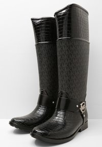 Bottes en caoutchouc noires avec une texture de crocodile sur les orteils et un dessus brillant. Le tige en tissu sombre arbore un motif de logo répété et des accents en métal.