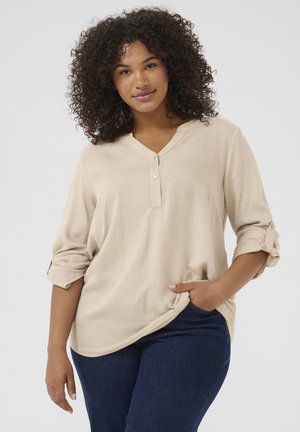 Camicia beige a maniche lunghe con scollo a V e patta con bottoni. Caratterizzata da polsini arrotolati e una vestibilità comoda, abbinata a jeans blu scuro.