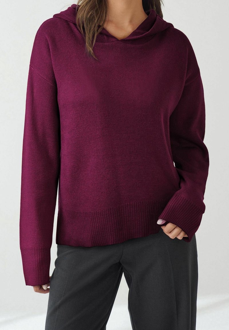 Femme portant un sweat à capuche en maille prune à manches longues associé à un pantalon ample noir, debout devant un fond clair uni.