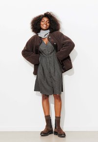 Veste en peau retournée marron portée sur une robe grise à boutons avec des pois, associée à des bottines marron à côtés élastiques et à des chaussettes texturées.