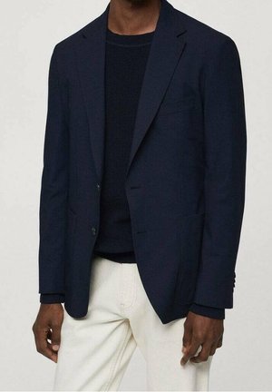 Giacca blazer blu navy con una vestibilità strutturata, risvolti a punta e due tasche frontali. Indossata sopra un maglione nero strutturato e pantaloni di colore chiaro.