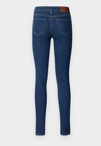 Jeans ajustados de color azul oscuro con dos bolsillos traseros, costuras visibles y una etiqueta de cuero marrón en la cinturilla, mostrados desde la parte trasera.