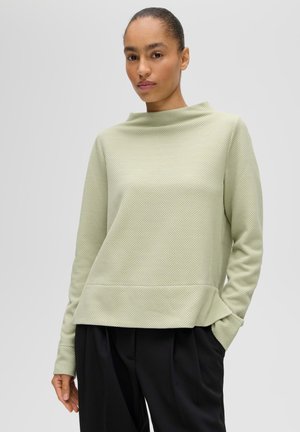 Donna con i capelli legati indossa una maglia a maniche lunghe verde chiaro con texture e pantaloni neri larghi, in piedi con una mano in tasca.