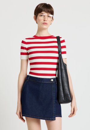 Donna con occhiali trasparenti, maglietta a righe rosse e bianche, gonna di denim scuro, che porta una borsa a tracolla nera su sfondo chiaro.