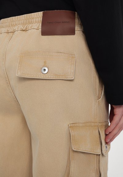 Beige cargo hlače iz bombaža z elastičnim pasom, ki imajo našitne žepe in usnjeno etiketo za nego z vtisnjenim besedilom.