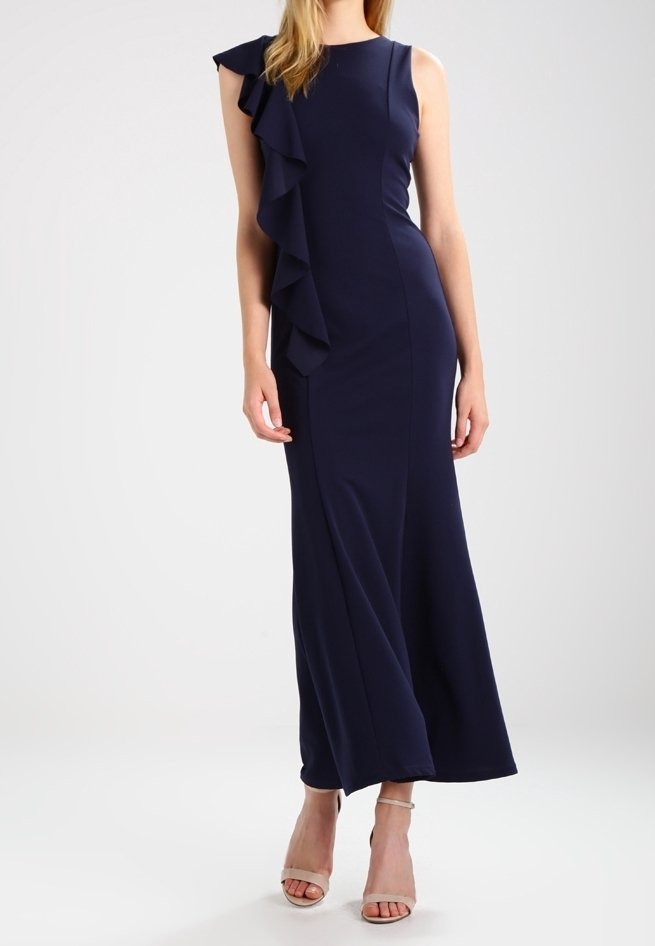 Femme portant une robe longue bleu marine sans manches avec un détail volanté sur une épaule et des sandales beiges à talons hauts.
