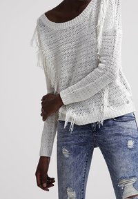 Weißer Strickpullover mit Fransenverzierung an den Seiten, rundem Ausschnitt und langen Ärmeln, kombiniert mit verwaschenen blauen Jeans.