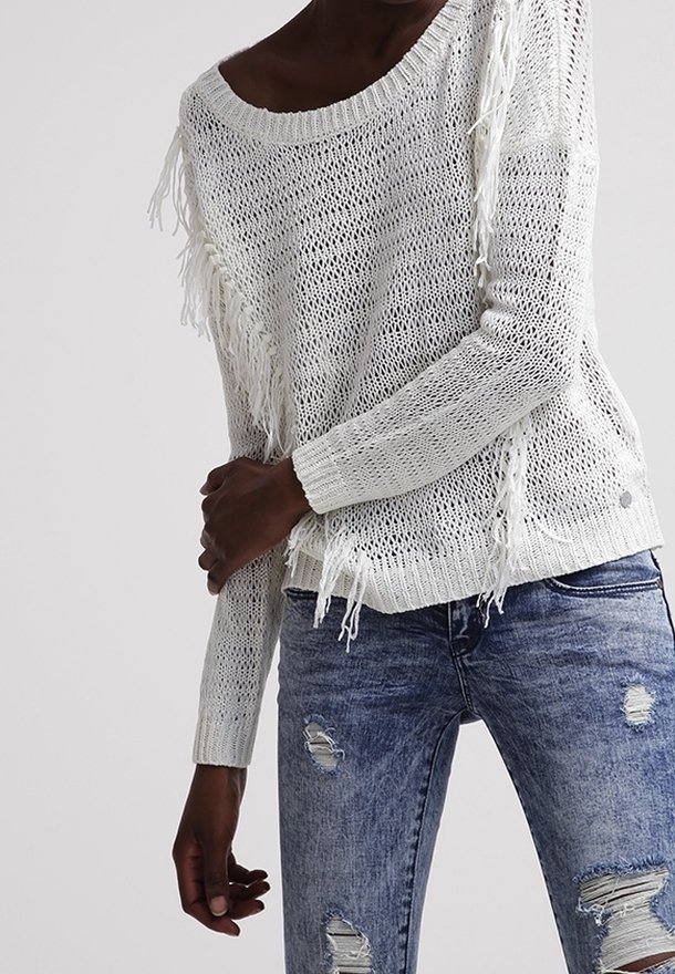 Weißer Strickpullover mit Fransenverzierung an den Seiten, rundem Ausschnitt und langen Ärmeln, kombiniert mit verwaschenen blauen Jeans.