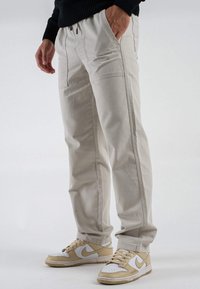 Jogging di un beige chiaro con vita elasticizzata e tasche laterali, caratterizzati da cuciture a contrasto. Abbinati a scarpe sportive beige e bianche.