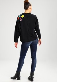Czarna bluzka z długim rękawem z kolorową haftowaną gwiazdką i zygzakiem, z zapięciem na guziki z tyłu. Połączona z dopasowanymi ciemnymi dżinsami i botkami za kostkę.