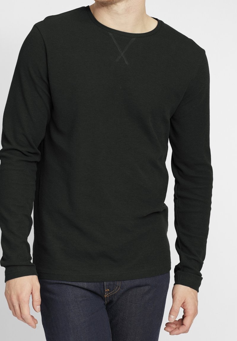 Homme portant une chemise noire à manches longues texturée et un jean bleu foncé, debout les bras détendus le long du corps devant un fond uni.