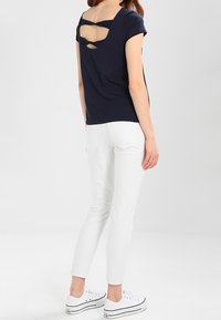 Kvinde iført en mørkeblå kortærmet top med crisscross rygdetalje, hvide slim-fit bukser og hvide platform sneakers, stående på hvidt gulv.