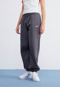 Sivé teplákové nohavice s elastickým pásom a manžetami, s bielym logom Nike na ľavom stehne. V páre s bielymi teniskami.