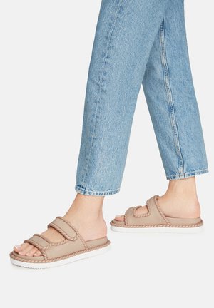 Svetlo bež natikači z drsno podlago, ki vključujejo teksturirano, oblazinjeno zgornjo stran z dvema nastavljivima trakoma in belo gumijasto podplat. Nosijo se z skrajšanimi kavbojkami.